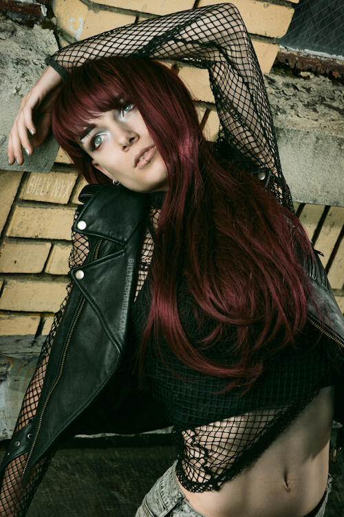Billy Nyx stretched out against a yellow brick wall with their arm stretched out and bent over their head. They have long red hair, a leather jacket, and a fishnet top on.